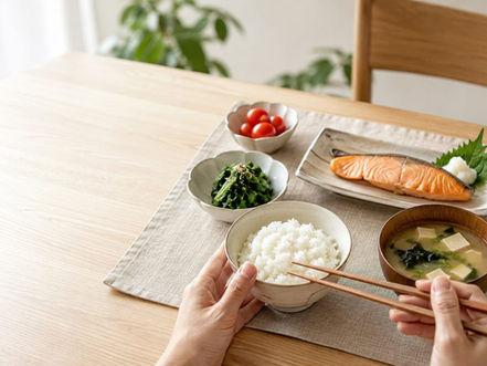 木のダイニングテーブルに白いごはん、焼き鮭、ほうれん草のおひたし、ミニトマト、豆腐とわかめの味噌汁が並び、手前で両手が茶碗と箸を持っている