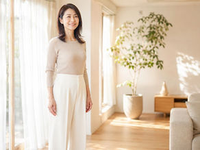 朝の光が差し込むナチュラルな雰囲気のリビングで、ベージュのリブニットとホワイトのワイドパンツを着た30代後半の女性が窓辺に立ち、穏やかに微笑んでいる様子