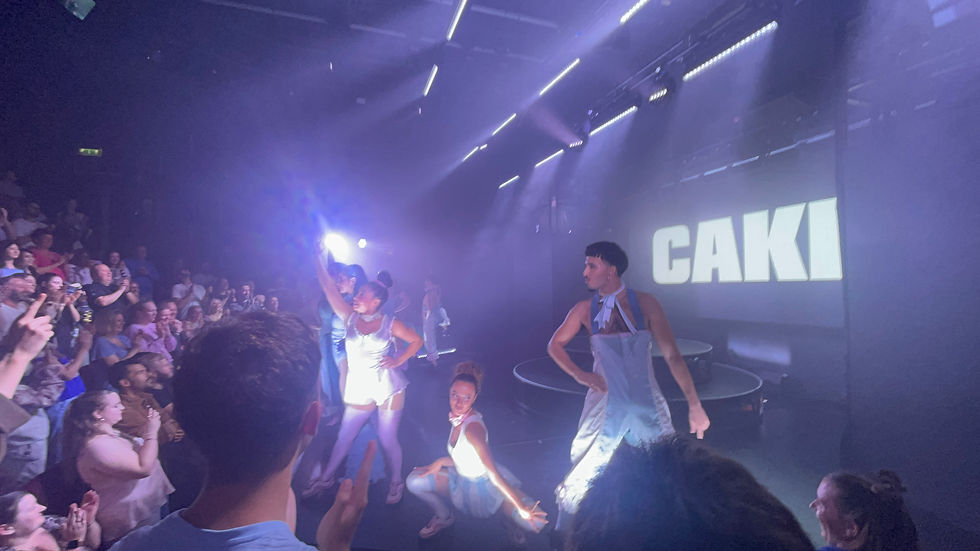 Performers in blue costumes dance on stage with a smoky ambiance. A crowd watches and applauds. "CAKE" is projected in bold letters.