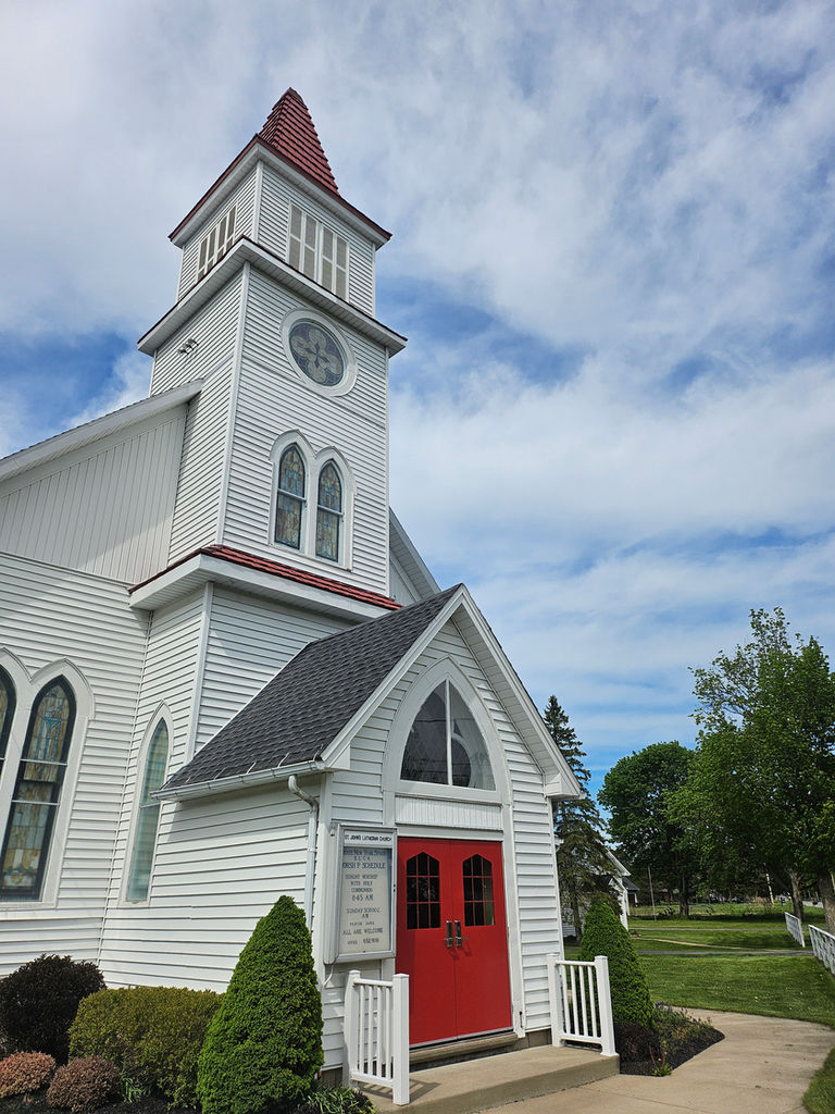 Exterior of St. Johns Lutheran Church with white siding, red doors, and steeple under a blue sky