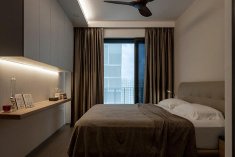 Small bedroom with white walls, study table, balcony door with sheer curtains, and dim lighting.

