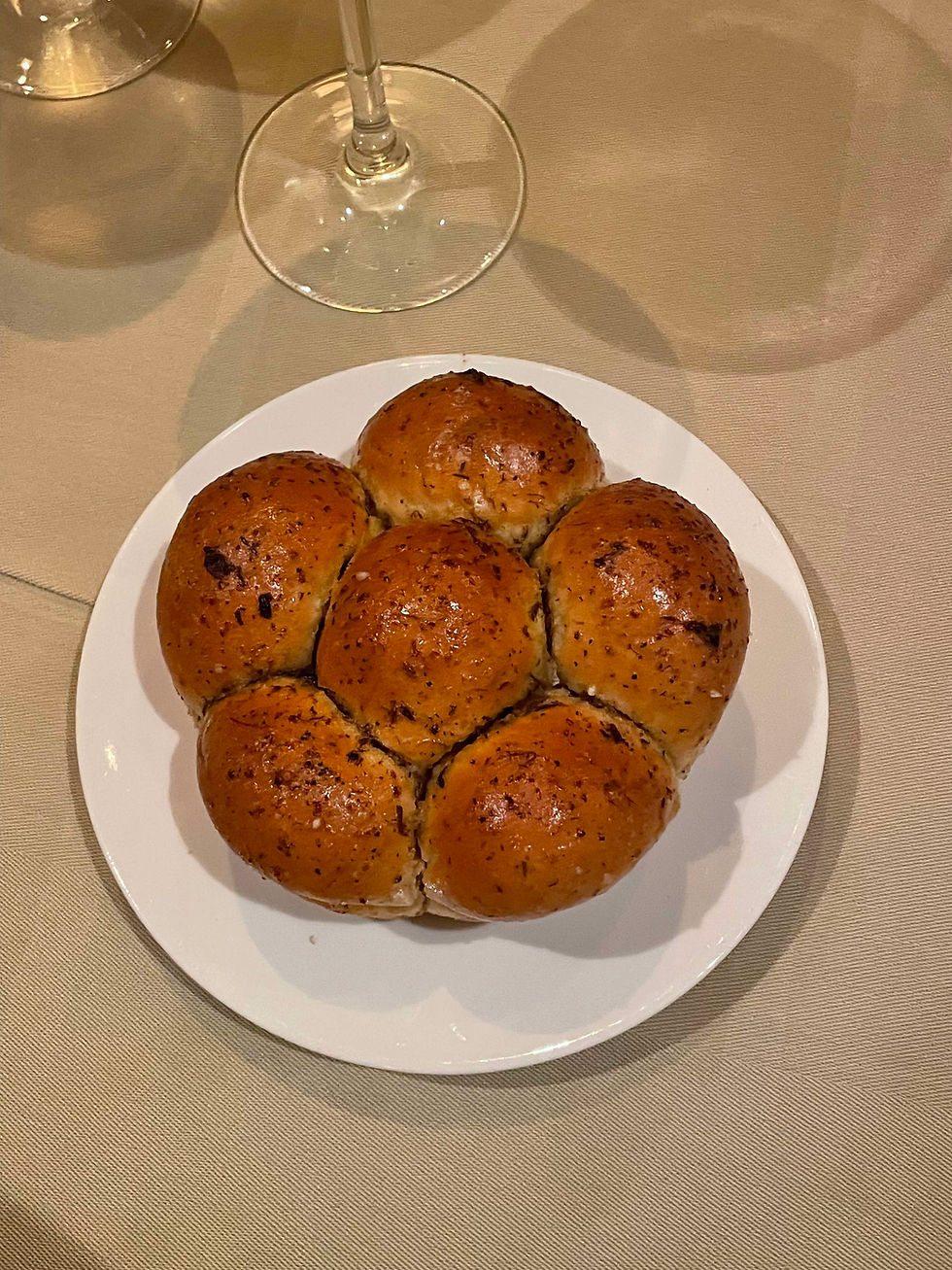 A close-up of the signature Parker House roll served at 150 Central Park aboard Utopia of the Seas. The golden-brown roll is brushed with truffle butter and topped with lemon pepper and thyme, sitting on a small white plate. Soft, pillowy, and glistening, the bread is plated simply—inviting, warm, and unforgettable.