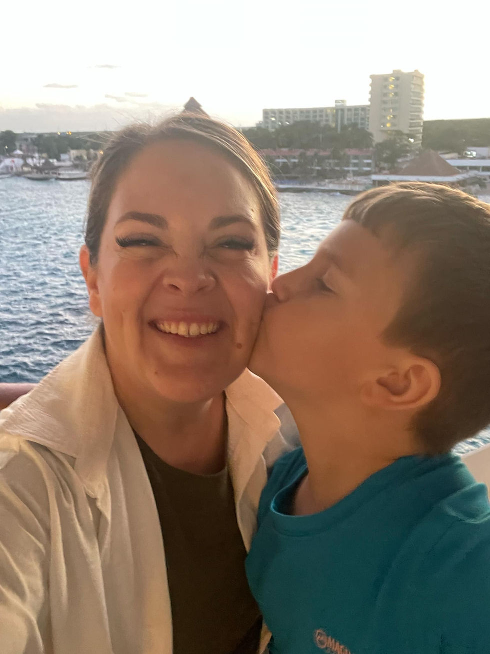 Selfie of travel agent Paige and her youngest son sharing a kiss on the cheek while standing on their oceanview balcony, overlooking the port of Cozumel during a January Royal Caribbean cruise.
