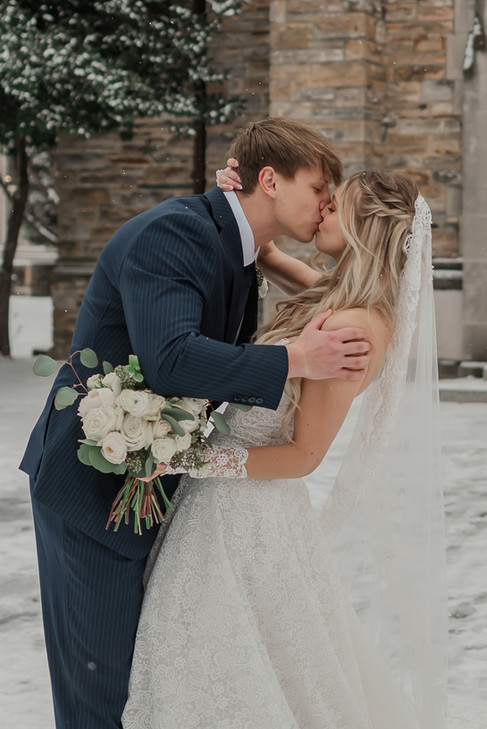 A bride and groom kiss outdoors in falling snow beside a stone building, with the bride holding a bouquet of white roses and greenery.