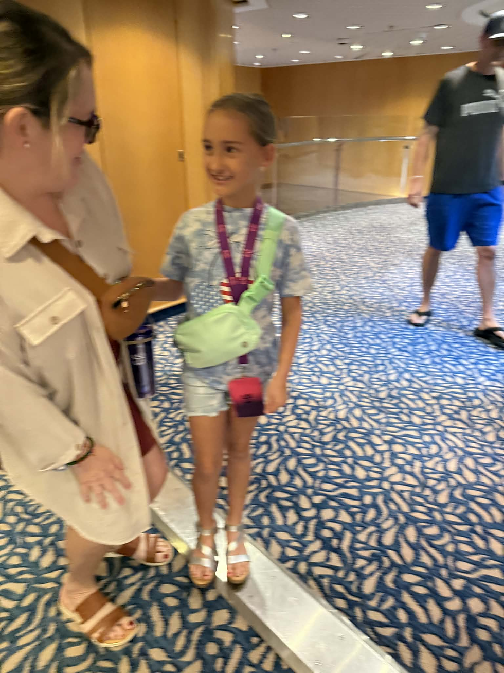 Woman and young girl smiling on a cruise ship, both wearing belt bags, with the girl holding a handmade bracelet featuring a small duck charm.