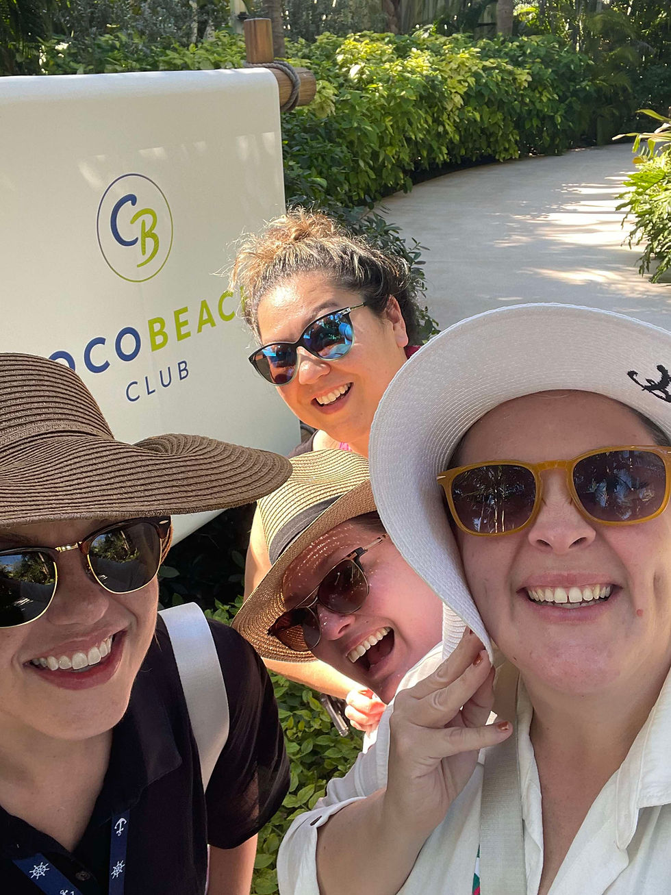 Four smiling women pose together at the entrance sign for Coco Beach Club on Royal Caribbean’s private island, Perfect Day at CocoCay. All are wearing sun hats or sunglasses and light beachwear, beaming in the sunshine. Lush greenery and a stone path frame the background, capturing the start of a luxurious, carefree day.