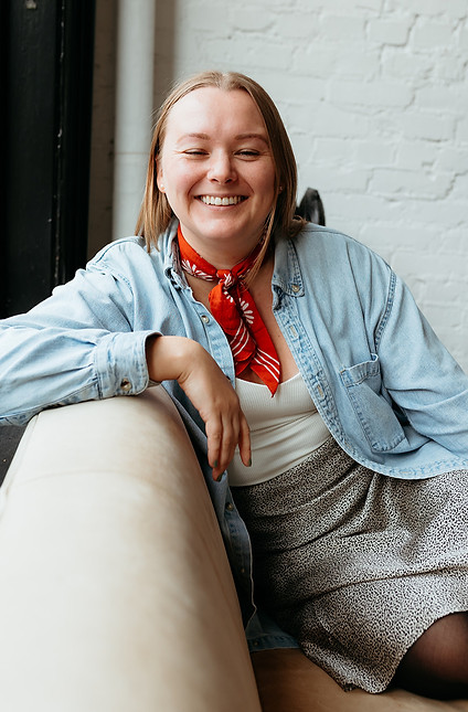 Woman with button up shirt and scarf sitting on a couch and smiling.