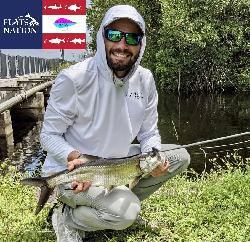 Smiling angler in white FLATS NATION hoodie kneels by a canal, holding a fish; logo overlays and mangroves in back.