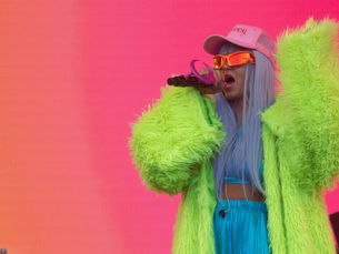 Li Saumet, chanteuse de Bomba Estéreo, sur scène au festival Fuego Fuego 2025 à Montréal. Elle porte un manteau vert fluo, un pantalon bleu métallisé et des lunettes orange, devant un fond rose éclatant.
