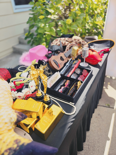 A prop table is filled with fun and exciting props including flower headbands, necklaces, a big stuffed fish, gold bars, oscar statues, sunglasses, hats, microphones, a phone shaped like lips, and a ukulele.