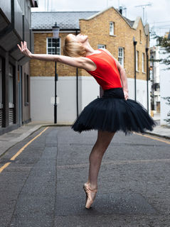 A ballerina in a red leotard and black tutu on pointe in the street