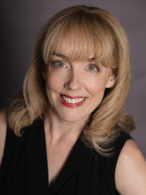 A headshot of a smiling blonde actress with a black top against a grey background