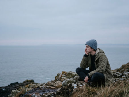 Uma pessoa solitária sentada de pernas cruzadas à beira de um penhasco rochoso, apoiando a cabeça na mão enquanto olha pensativamente para o mar sob um céu cinzento e nublado.