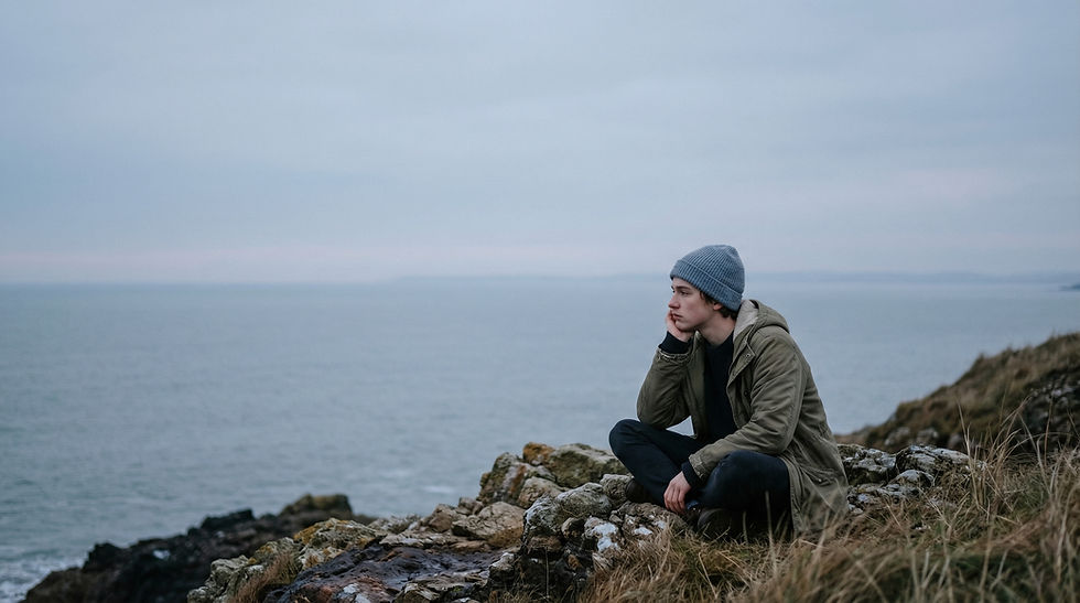 Uma pessoa solitária sentada de pernas cruzadas à beira de um penhasco rochoso, apoiando a cabeça na mão enquanto olha pensativamente para o mar sob um céu cinzento e nublado.