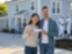 Smiling couple holds a check in front of a gray house. Painters work in the background. Bright day, joyful mood.