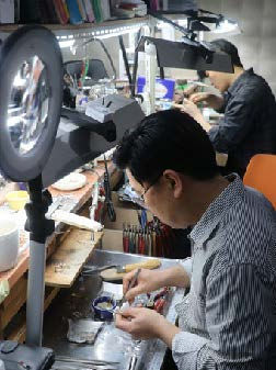 Two jewelers work meticulously at a cluttered workbench under bright lamps. The room is filled with tools and materials, creating a focused mood.