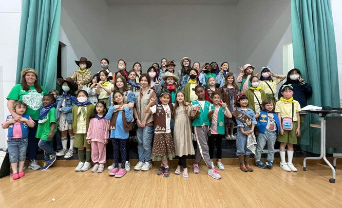 A large group of children in colorful scout uniforms pose smiling on a stage with green curtains in a hall, some wearing hats or masks.