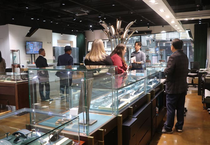 People in a jewelry store converse around glass display cases with watches, set in a modern interior. Neutral tones and overhead lighting.