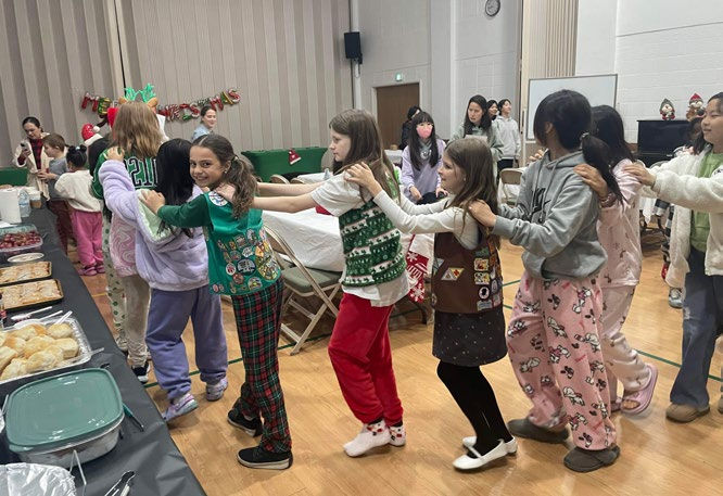 Children in festive clothing form a conga line at a holiday party. Tables with food and decorations are in the background. Joyful mood.