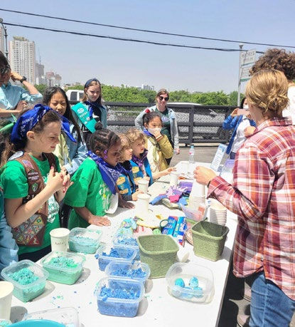 Children in green outfits gather around a table with blue craft materials outdoors, guided by an adult in a plaid shirt, on a sunny day.