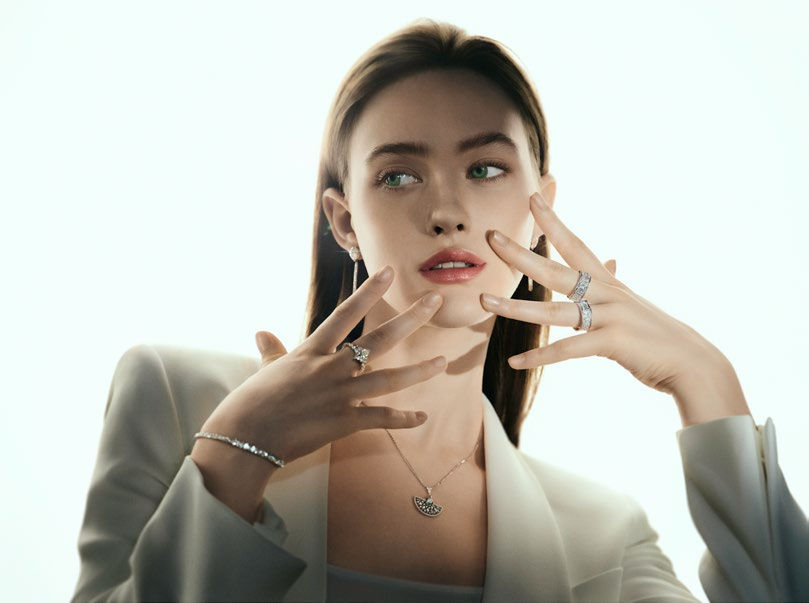 Woman in white suit, displaying jewelry with hands. Background is bright and minimal. Mood is elegant and contemplative. Photo courtesy of Aldo
