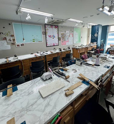 Empty workshop with long white tables, tools scattered around, and wooden chairs. Boards with notes and decorations on walls. Bright lighting.