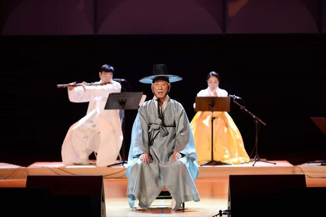 Musicians in traditional attire perform on stage; a seated man sings, flanked by a flutist and a woman in a yellow dress behind.