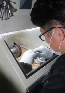 Person wearing gloves and a mask works on machinery inside a glass-enclosed workspace. Tools hang on the wall in a workshop setting.