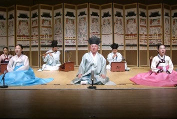 Performers in traditional Korean attire sing on stage, wearing pastel colors. A decorative folding screen serves as the backdrop.