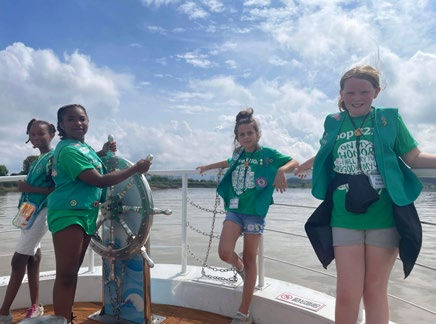 Four girls in green shirts on a boat, one steering. Smiling under a partly cloudy sky, with water in the background. Bright, cheerful scene.