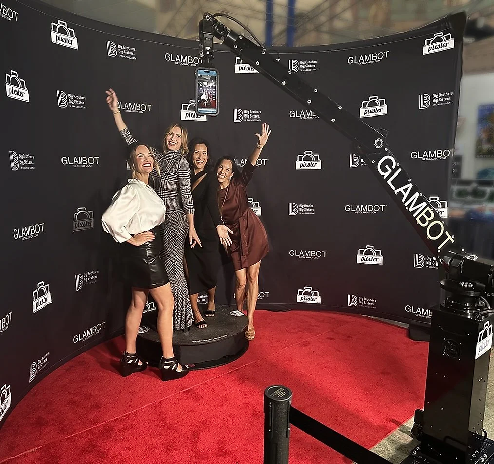 Four women pose joyfully on a red carpet in front of a GLAMBOT backdrop, under a camera crane. They're dressed stylishly, exuding excitement. By DMA Events