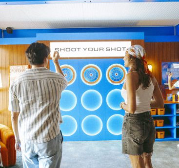 Two festival attendees enjoy an interactive "Shoot Your Shot" digital game at a vibrant DMA Coachella brand activation, featuring a sleek blue and wood-toned lounge design with modern festival branding.
