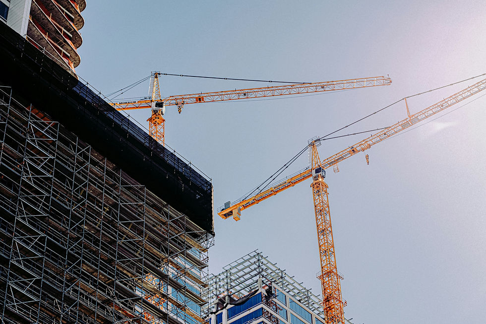 Two yellow cranes over construction site with scaffolding and buildings against a clear blue sky. Sunlight creates a bright atmosphere.