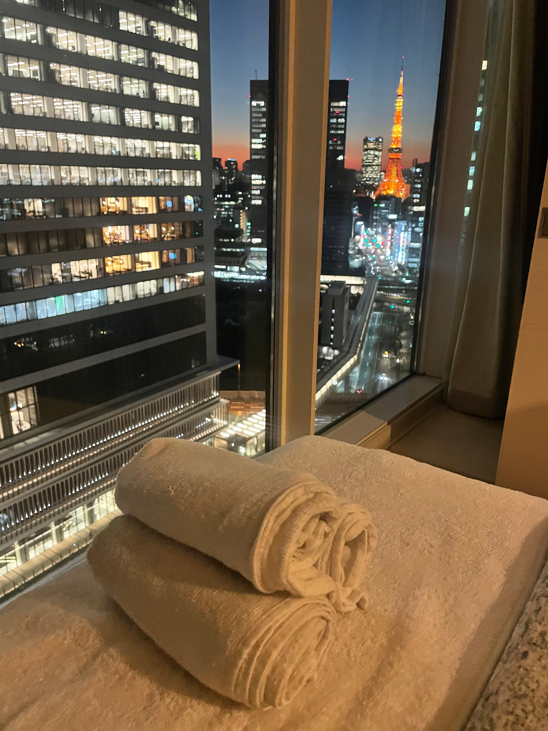 A treatment bed in front of a large window, with the dusk skyline of Tokyo and Tokyo Tower glowing in the background