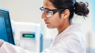 A female scientist wearing a white lab coat, safety glasses, and gloves as she interacts with a digital computer monitor displaying laboratory or diagnostic data.