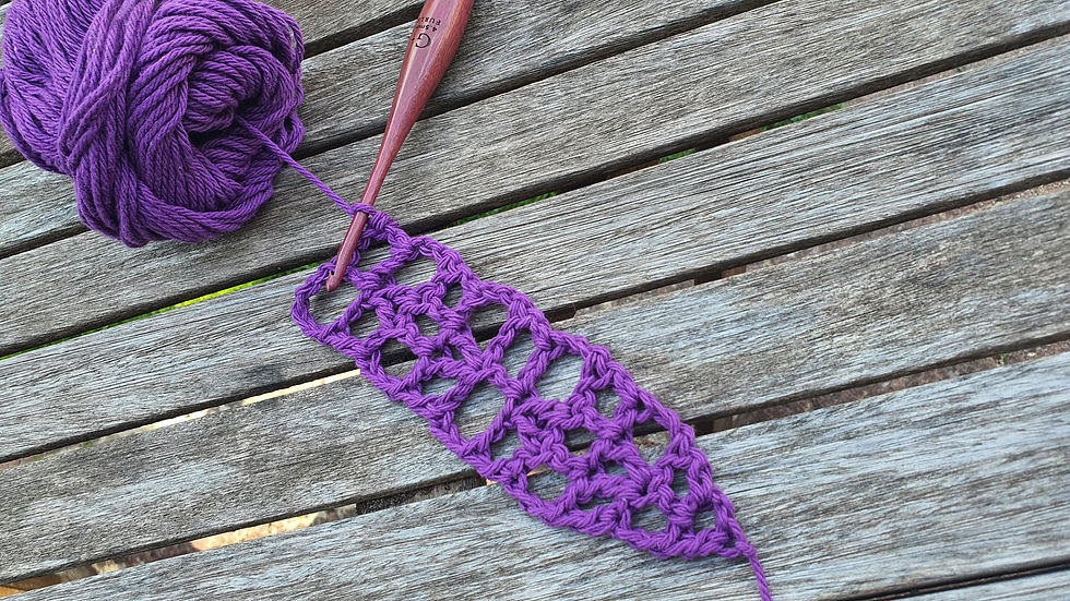 Purple yarn and a wooden crochet hook working on an open lace pattern on a weathered wooden table. The mood is calm and creative.