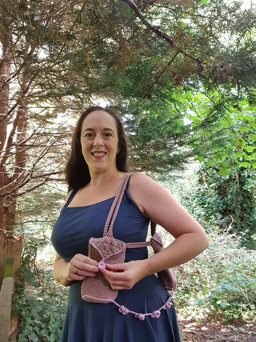 Smiling woman in a blue dress holds a brown and pink crocheted bag. She stands in a lush, green forest with sunlight filtering through the trees.