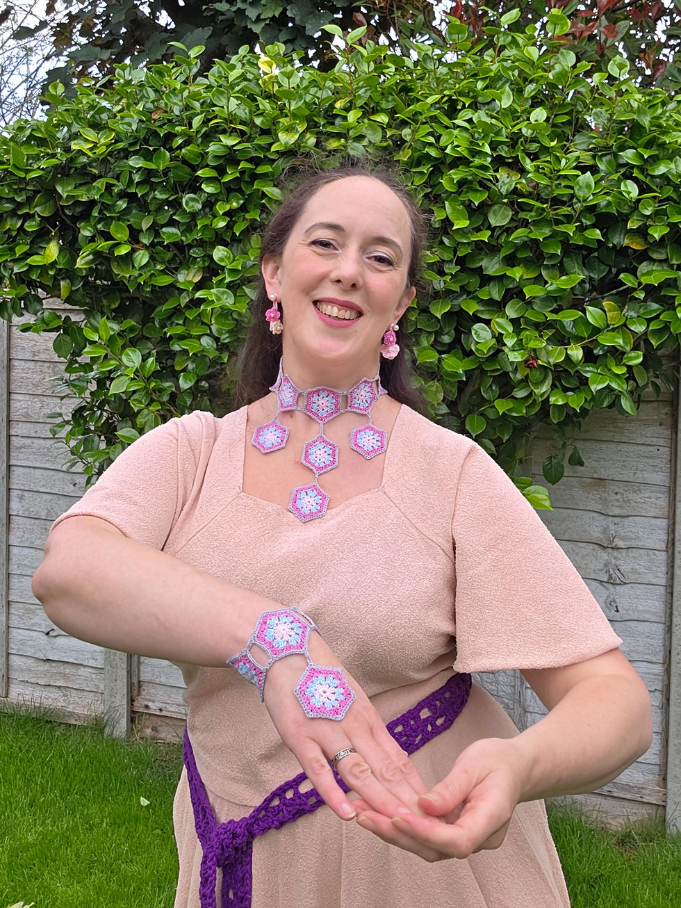 Smiling woman in a garden, wearing pink dress and crocheted hexagon jewelry, posing before a leafy hedge.