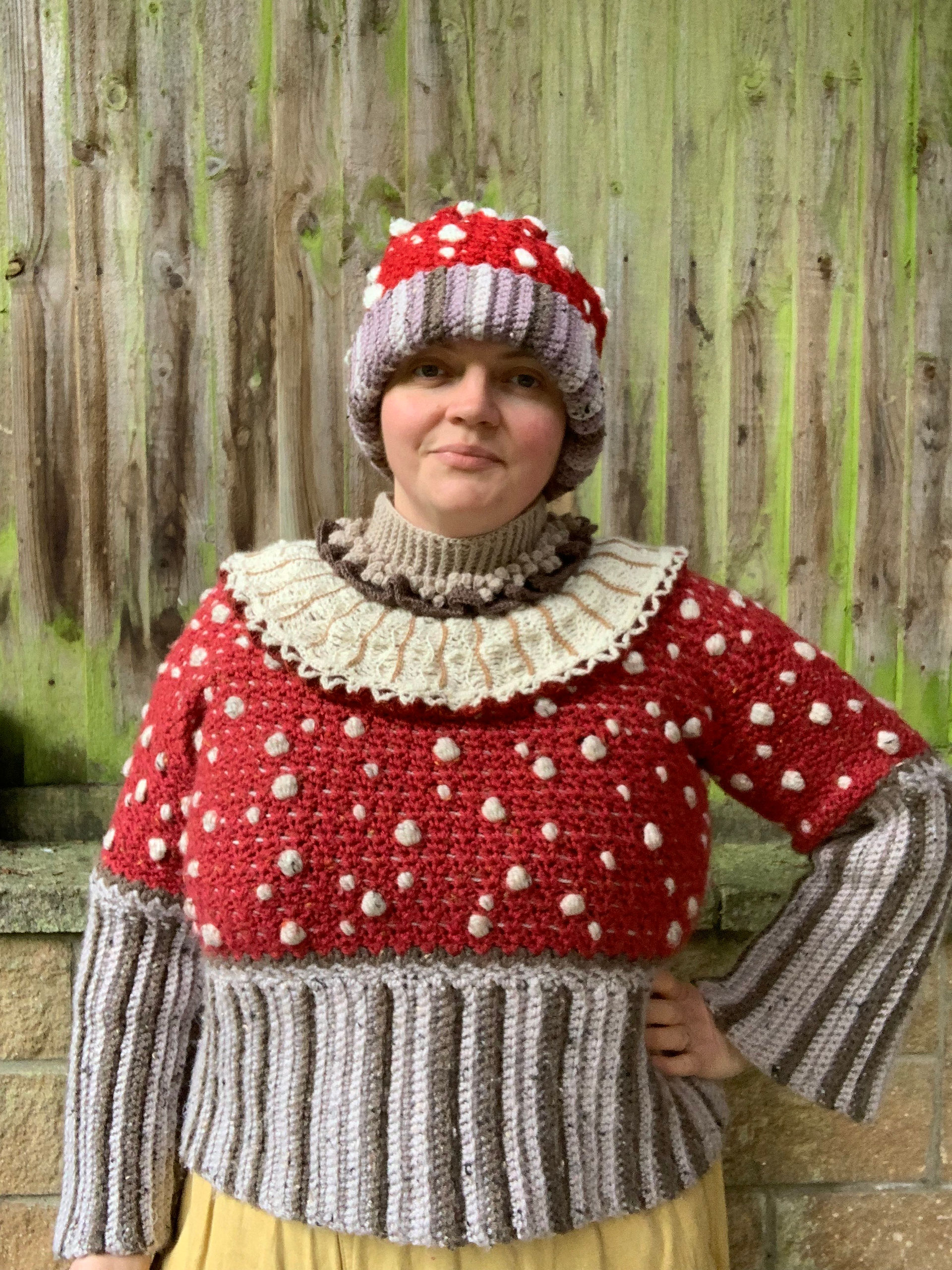 Woman wearing a cream and brown crocheted yoked cowl teamed with other crocheted clothing against a wooden fence