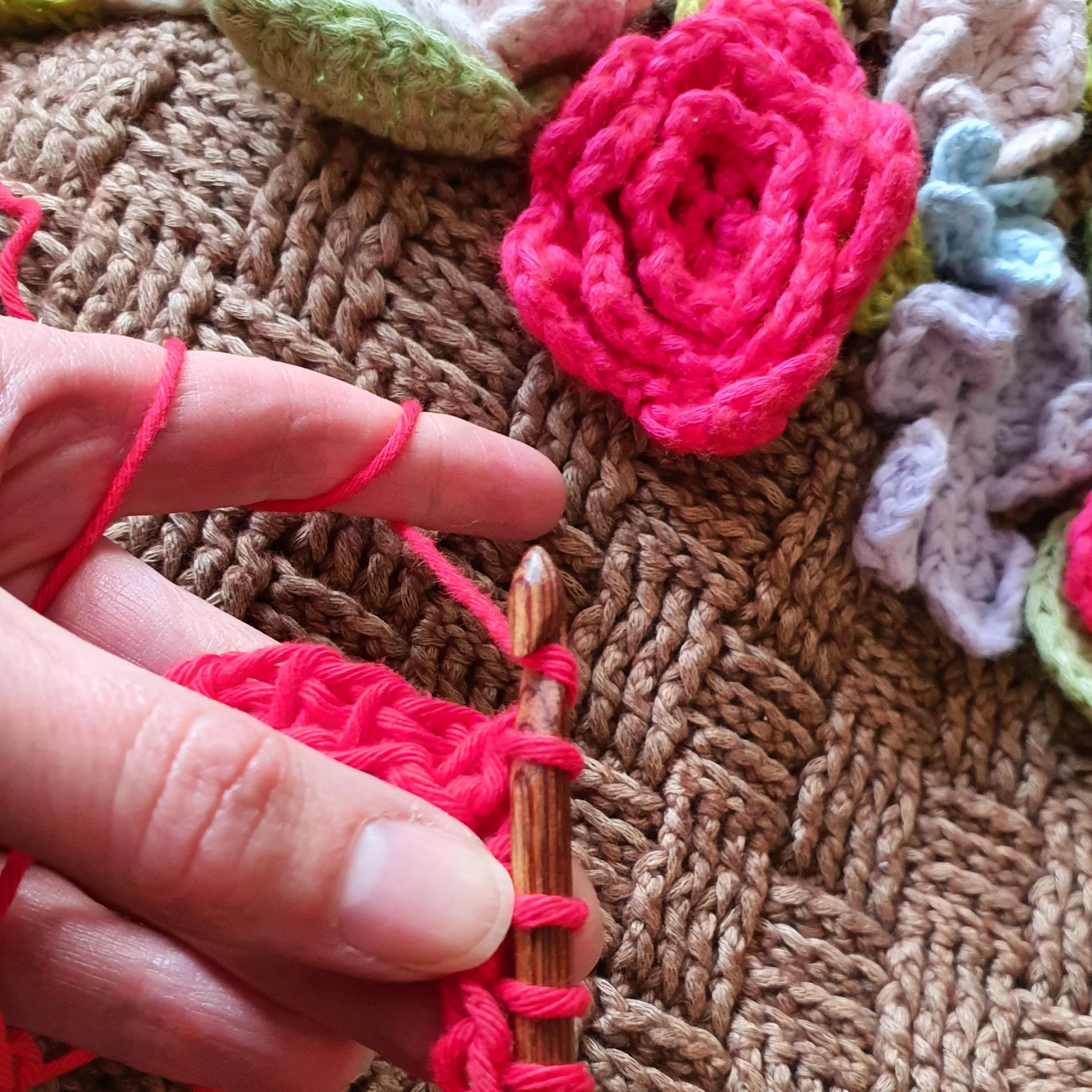 The Flower Basket Wrap is a brown cotton shawl crocheted in basketweave stitch and lattice stitch with a border at either end covered with brightly coloured crocheted flowers in a range of styles and shades. It's shown in use over a table with hands crocheting in front