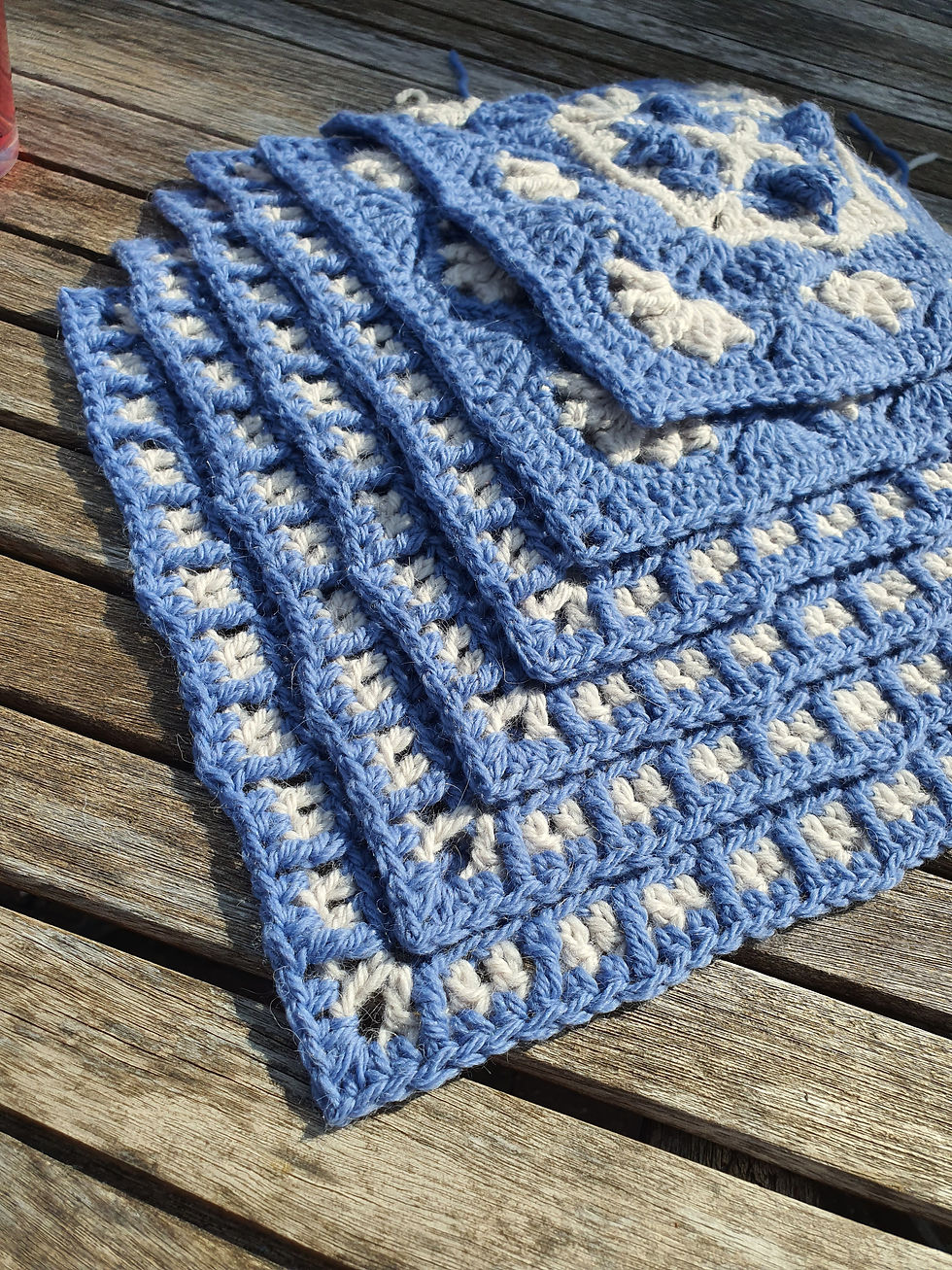 Stack of blue and greycrochet squares on a wooden table, showcasing intricate patterns in bright sunlight.