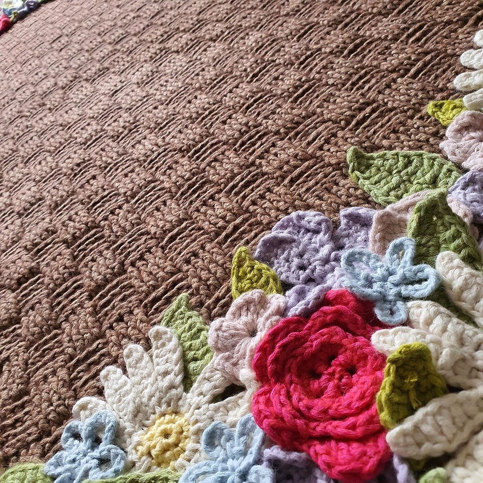 The Flower Basket Wrap is a brown cotton shawl crocheted in basketweave stitch and lattice stitch with a border at either end covered with brightly coloured crocheted flowers in a range of styles and shades. It's shown close to one end of the border with the other end in the distance