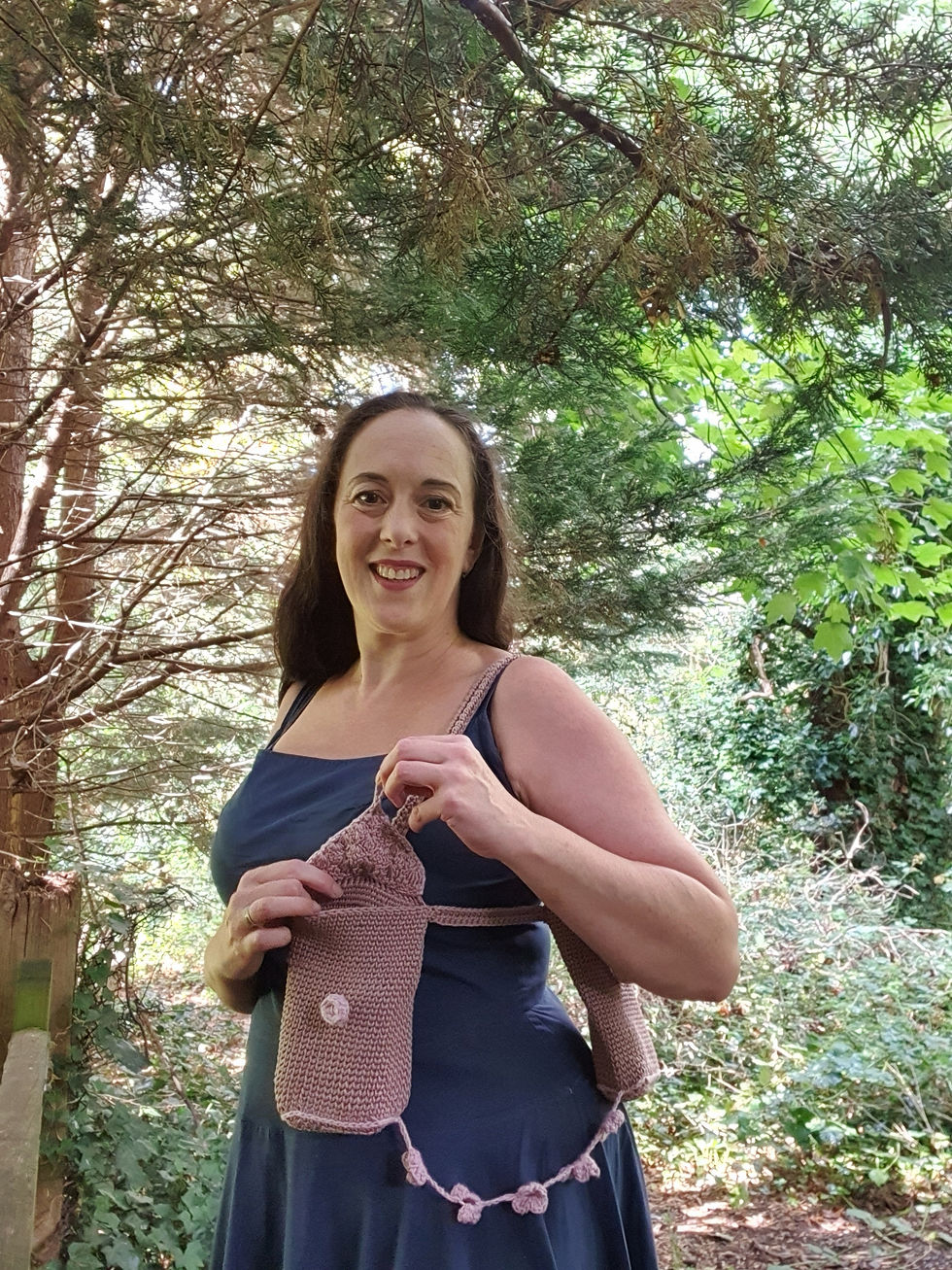 Woman in a blue dress stands in a forest, smiling and holding a brown and pink crocheted bag. Sunlight filters through green leaves.