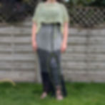 Woman in a crocheted grey and black skirt and green top stands on grass near a wooden fence and leafy bushes, under a clear sky.