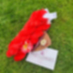 Coral yarn and a pink flower sit on grass with a crochet hook, wooden dish, tape measure, and "Miss Babs" card. Bright and vibrant scene.