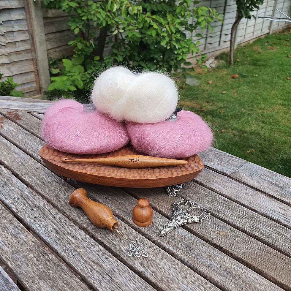 Pink and white fluffy yarn balls, crochet hook, and tools on a wooden table outdoors. Greenery in the background. Relaxed crafting vibe.