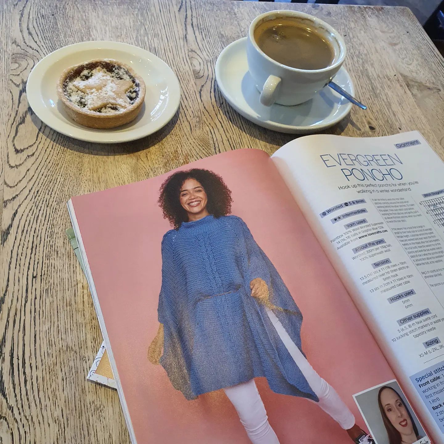 A crochet magazine featuring a blue cabled poncho on a wooden table with coffee and a mince pie