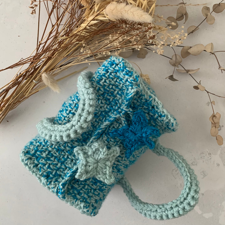 The Amelina Small Project Bag is a rectangular bag with square ends, shown made in blue and turquoise variegated yarn with dried flowers behind