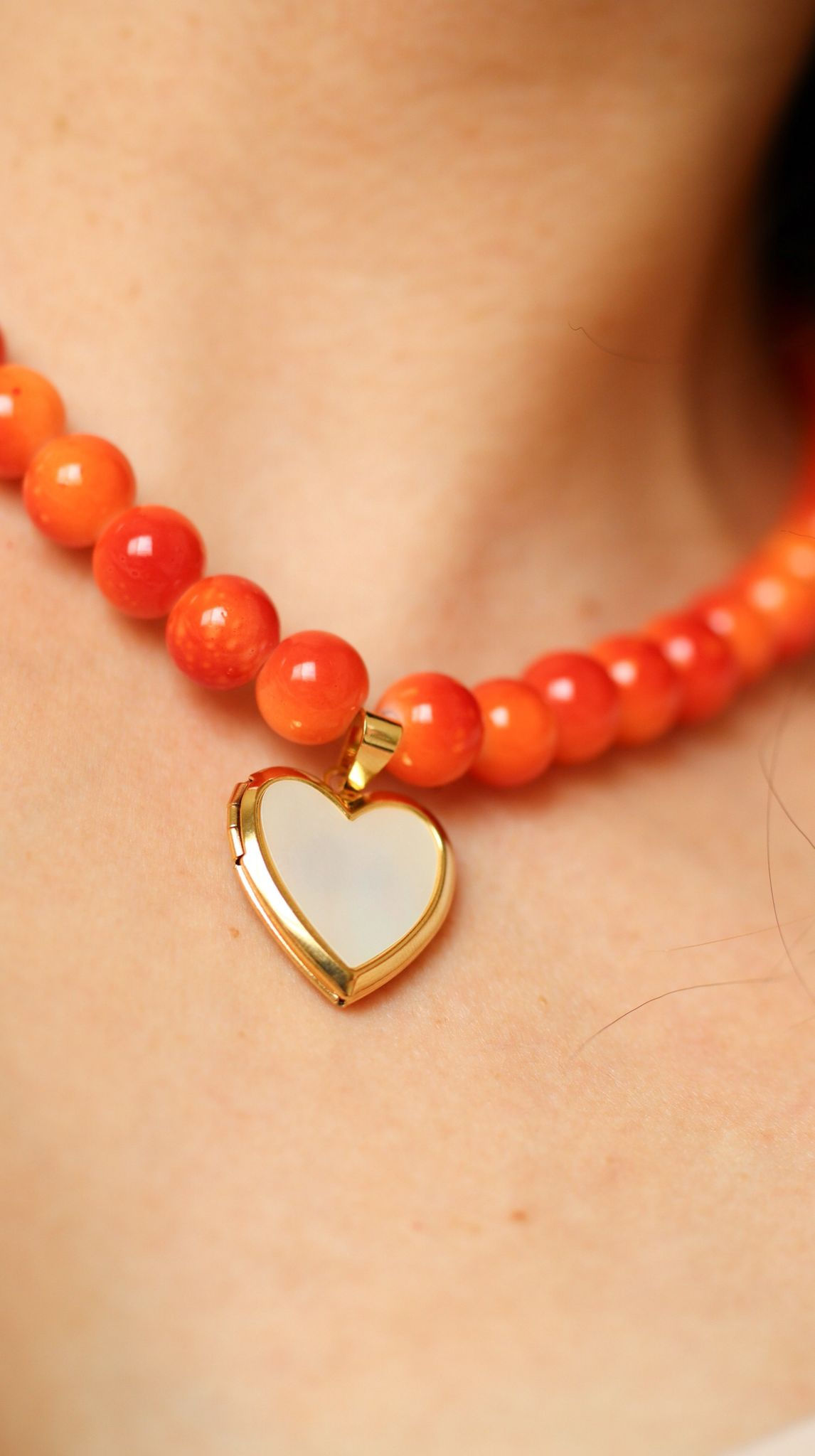 Close-up of orange glass bead necklace with heart-shaped pearl locket and gold stainless steel details.