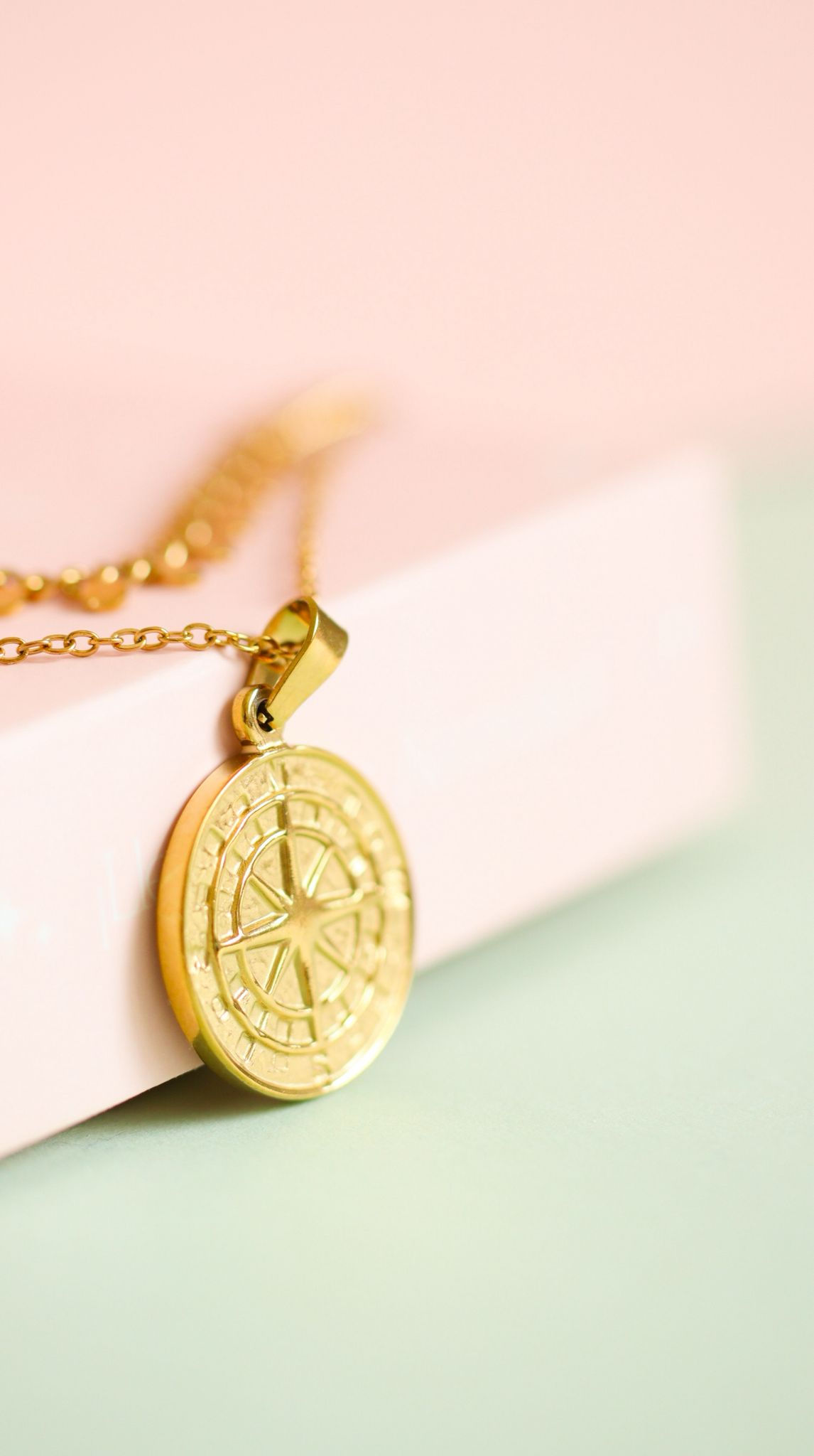 Close-up of gold stainless steel compass pendant necklace displayed on soft pink background.